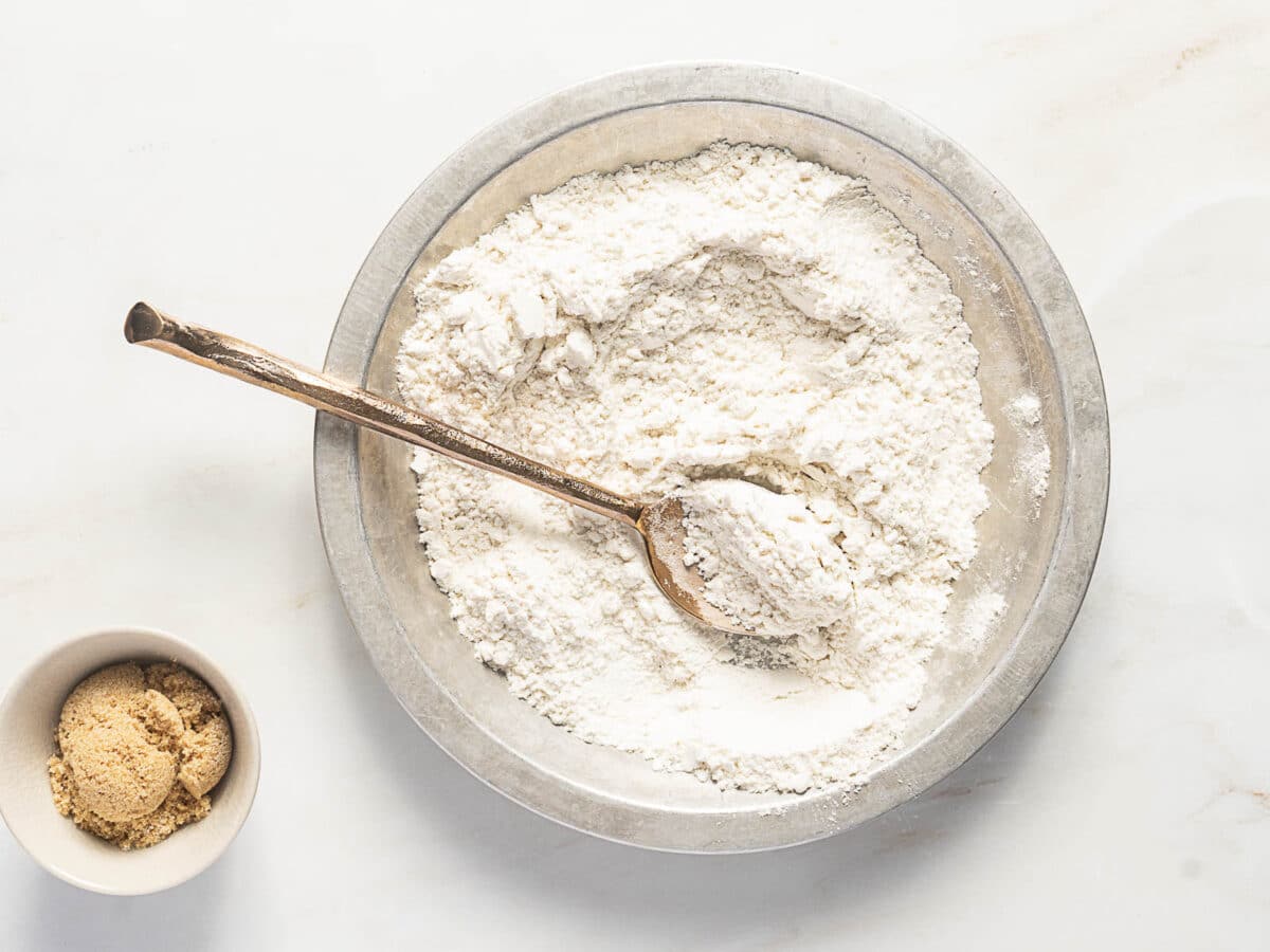A metal bowl filled with flour and a metal spoon sits on a white surface; a small bowl of brown sugar is next to it.