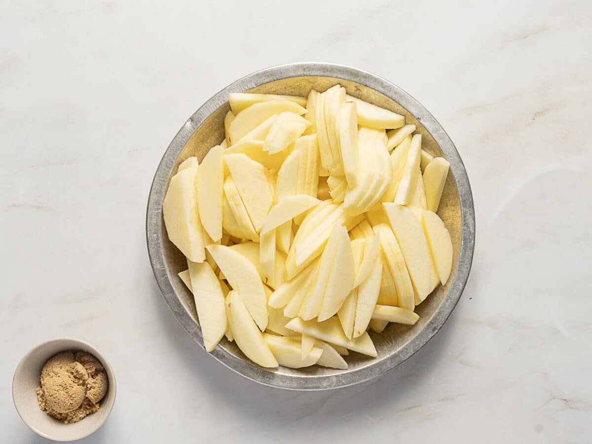 A metal bowl filled with sliced apples sits on a marble surface next to a small bowl containing brown sugar.
