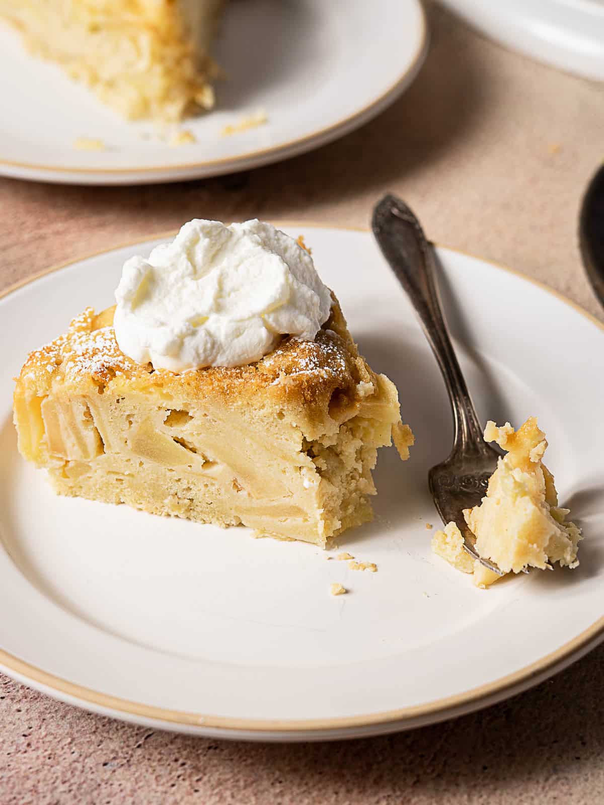 A slice of apple cake topped with whipped cream sits on a white plate next to a fork with a bite of cake.