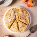 A round apple pie with lattice crust is dusted with powdered sugar and partially sliced, placed on a white plate beside apples, plates, and a pie server.
