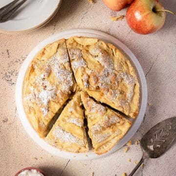 A round apple pie with lattice crust is dusted with powdered sugar and partially sliced, placed on a white plate beside apples, plates, and a pie server.