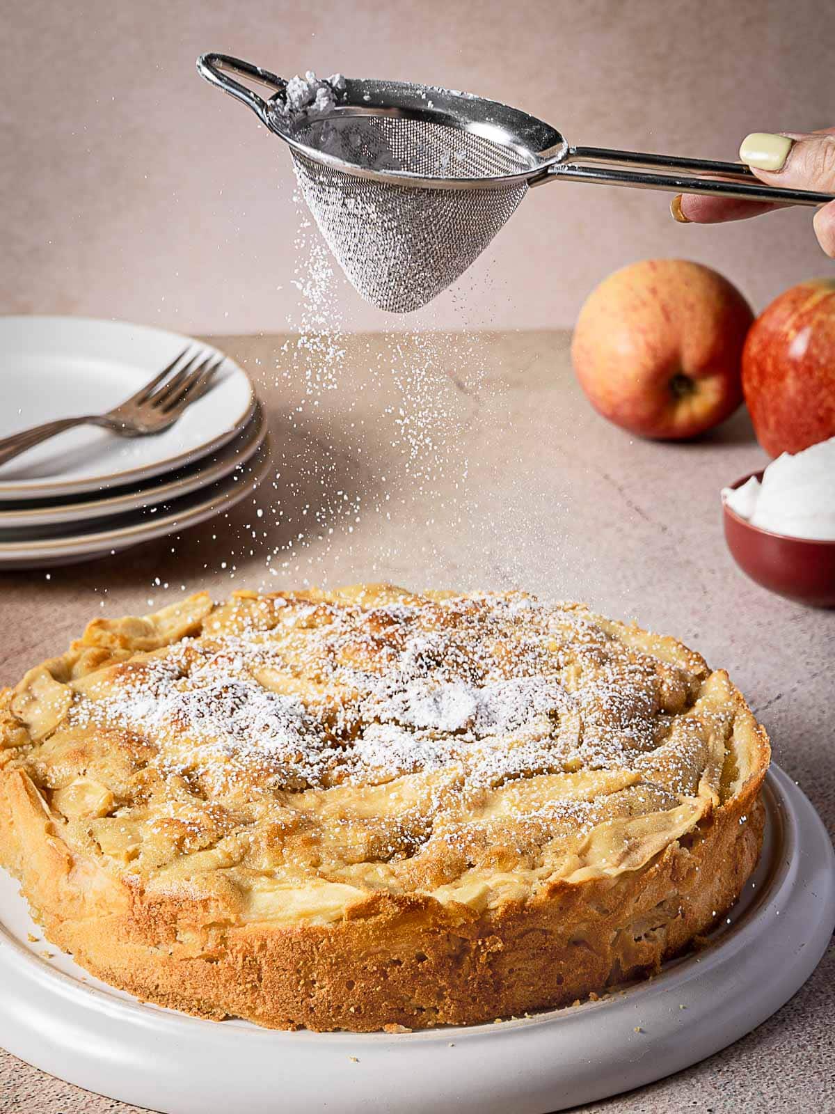 A hand sifts powdered sugar over a baked apple cake on a white plate, with apples, plates, and a bowl of whipped cream in the background.