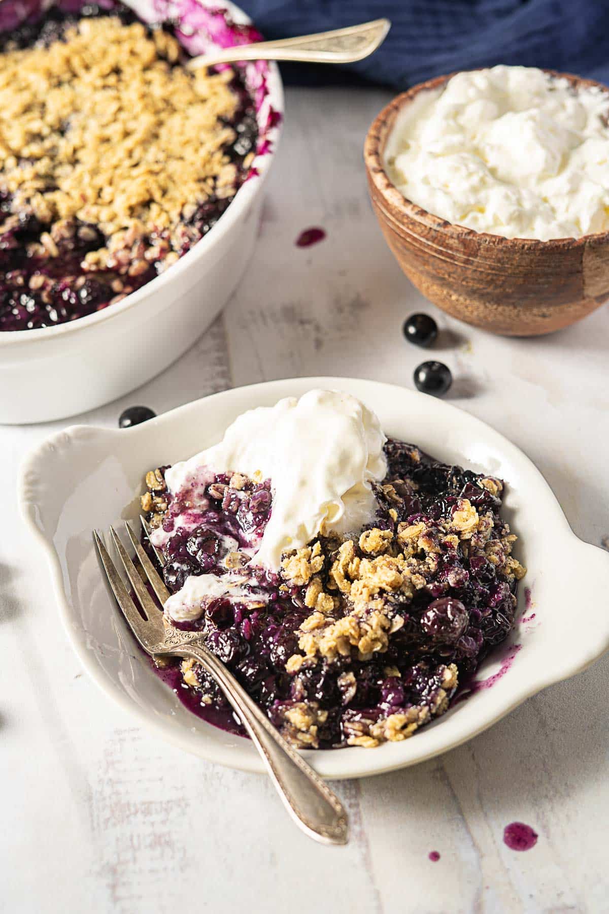 A plate of blueberry crisp topped with a scoop of vanilla ice cream, with a fork, next to a bowl of whipped cream and a baking dish of blueberry crisp.