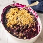 A baked blueberry crisp in a white oval dish, topped with a golden oat crumble, with a serving spoon and some filling missing from one side.