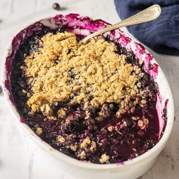 A baked blueberry crisp in a white oval dish, topped with a golden oat crumble, with a serving spoon and some filling missing from one side.