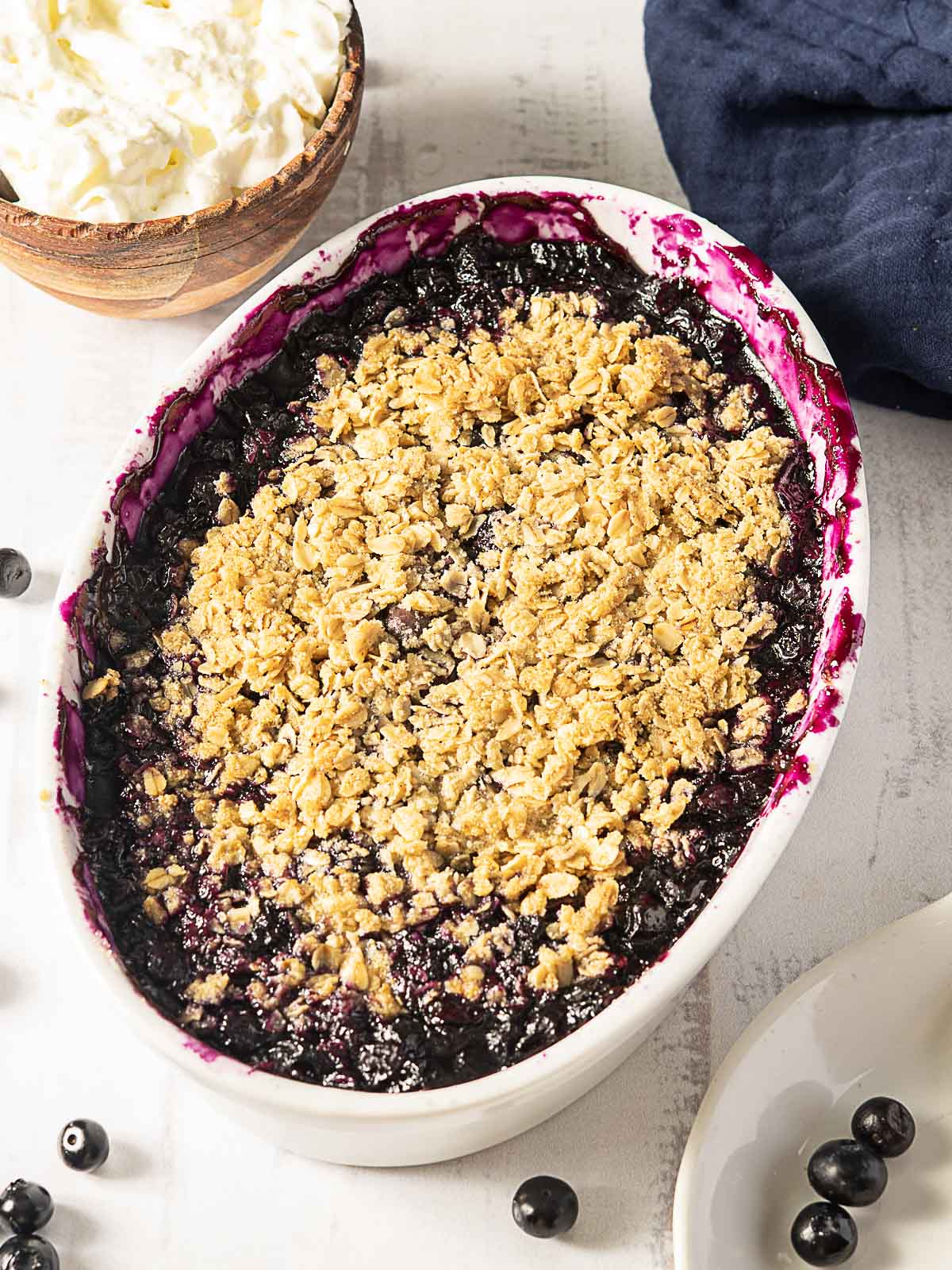 An oval baking dish filled with blueberry crisp topped with a golden oat crumble, surrounded by fresh blueberries, a bowl of whipped cream, and a navy cloth.
