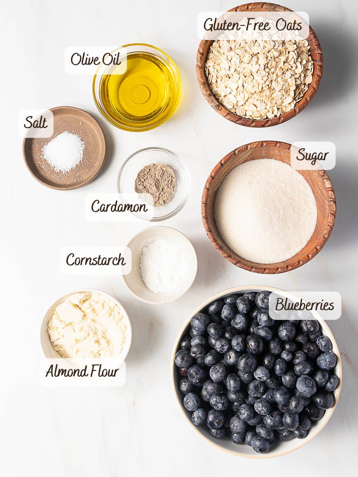 A flat lay of ingredients for a Gluten Free Blueberry Crisp, including gluten-free oats, olive oil, salt, cardamom, sugar, cornstarch, almond flour, and blueberries, each labeled in small bowls.