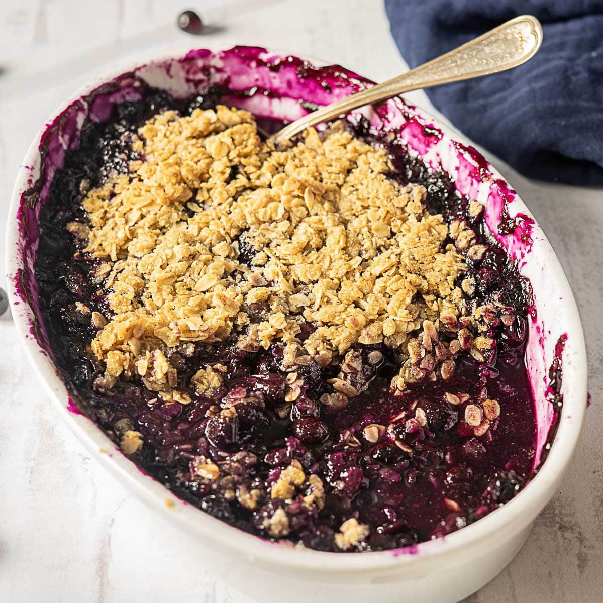 A baked blueberry crisp in a white oval dish, topped with a golden oat crumble, with a serving spoon and some filling missing from one side.