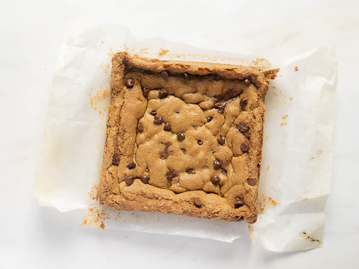 A baked square chocolate chip blondie sits on crumpled parchment paper atop a white surface, with crumbs scattered around the edges.
