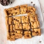 A batch of chocolate chip cookie bars cut into squares on parchment paper, with a small bowl of chocolate chips nearby.
