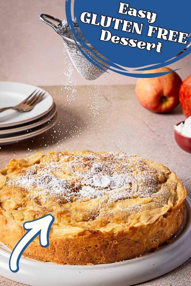 A round gluten free apple cake is dusted with powdered sugar. Plates, apples, and a bowl of sugar are in the background. Text reads "Easy GLUTEN FREE Dessert.