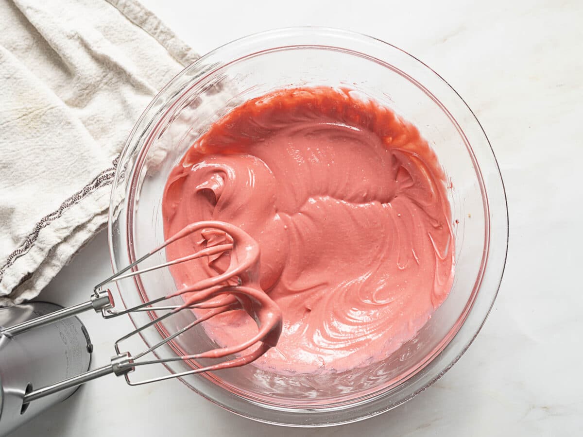A glass mixing bowl filled with pink batter and an electric hand mixer with beaters, on a white countertop beside a cloth.