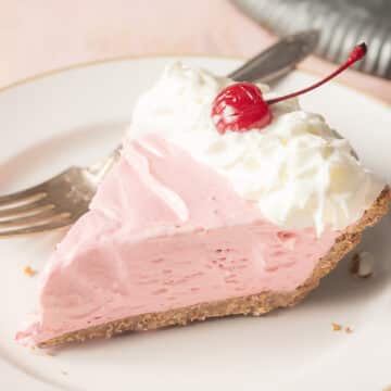 A slice of pink pie with a graham cracker crust, topped with whipped cream and a maraschino cherry on a white plate with a fork.