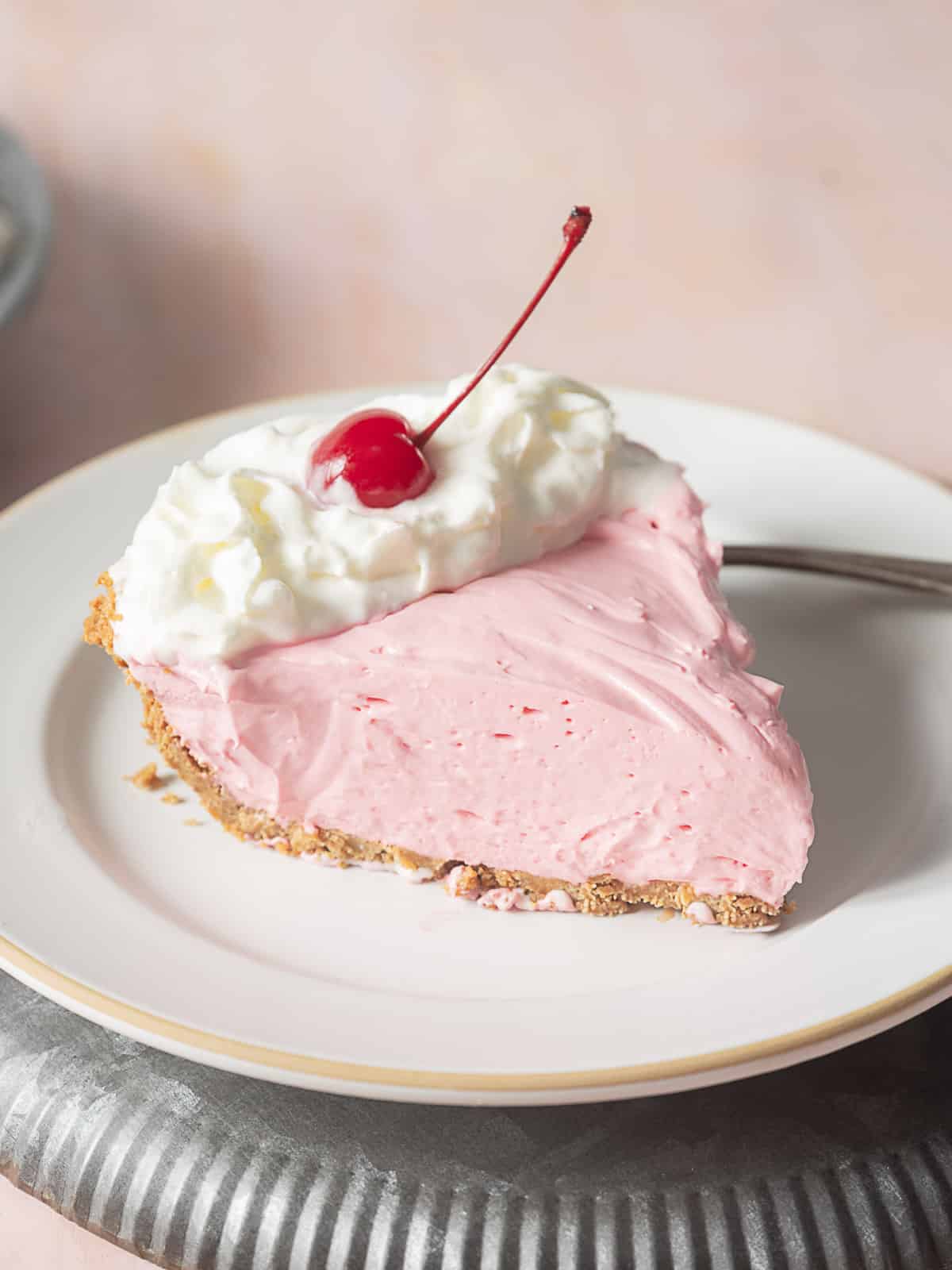 A slice of pink cream pie with a graham cracker crust, topped with whipped cream and a cherry, on a white plate.