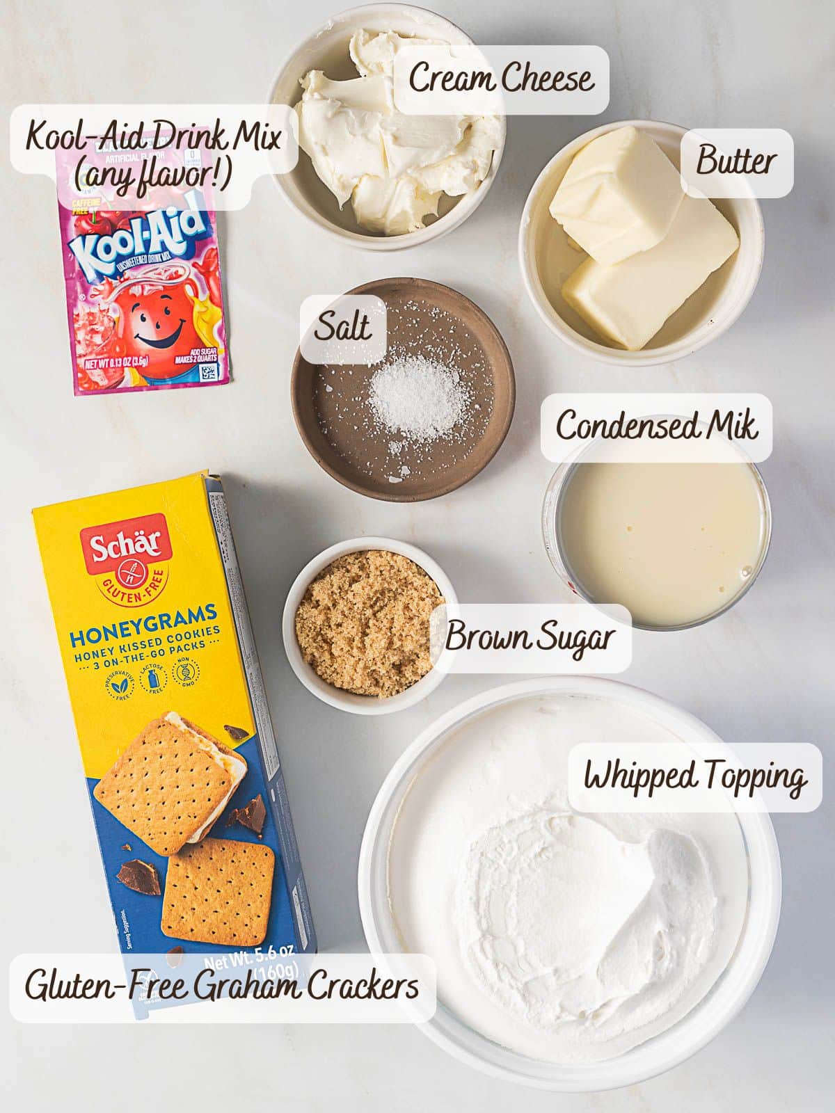 Ingredients for a gluten free dessert no bake treat are laid out, including Kool-Aid drink mix, cream cheese, butter, salt, condensed milk, brown sugar, whipped topping, and gluten-free graham crackers.
