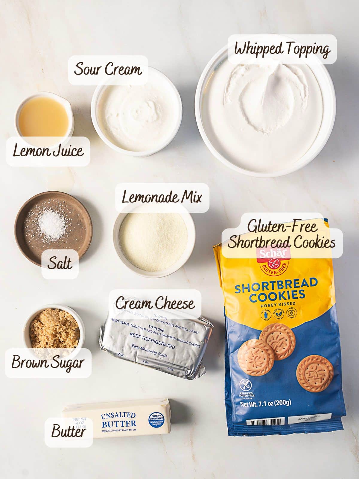 Ingredients for a gluten free lemon pie are laid out on the counter: sour cream, lemon juice, salt, lemonade mix, whipped topping, gluten-free shortbread cookies, brown sugar, cream cheese, and butter.