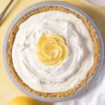 A lemon cream pie with a graham cracker crust, topped with whipped cream and lemon slices, sits on a light surface next to a whole lemon.