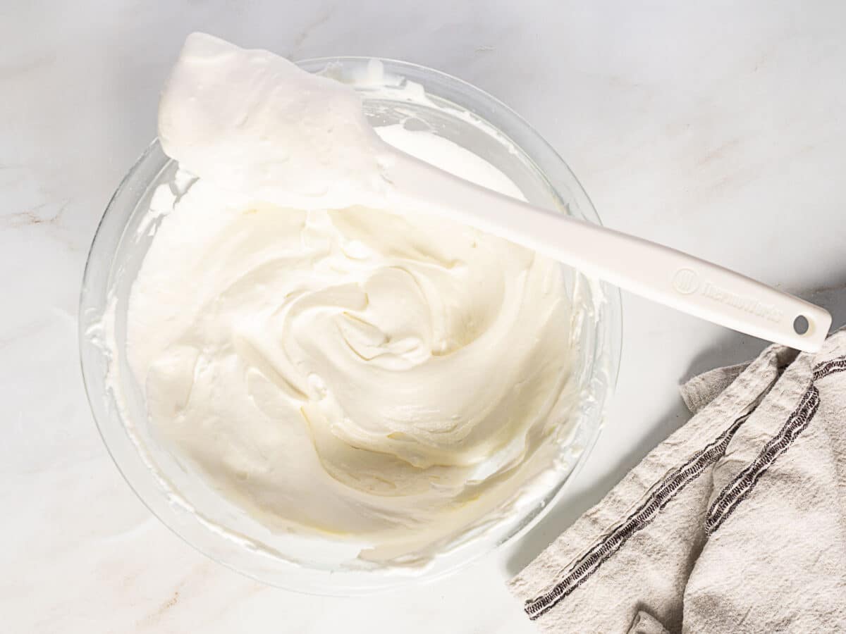 A glass bowl filled with whipped cream and a white spatula resting across the top, next to a folded beige kitchen towel on a light surface.