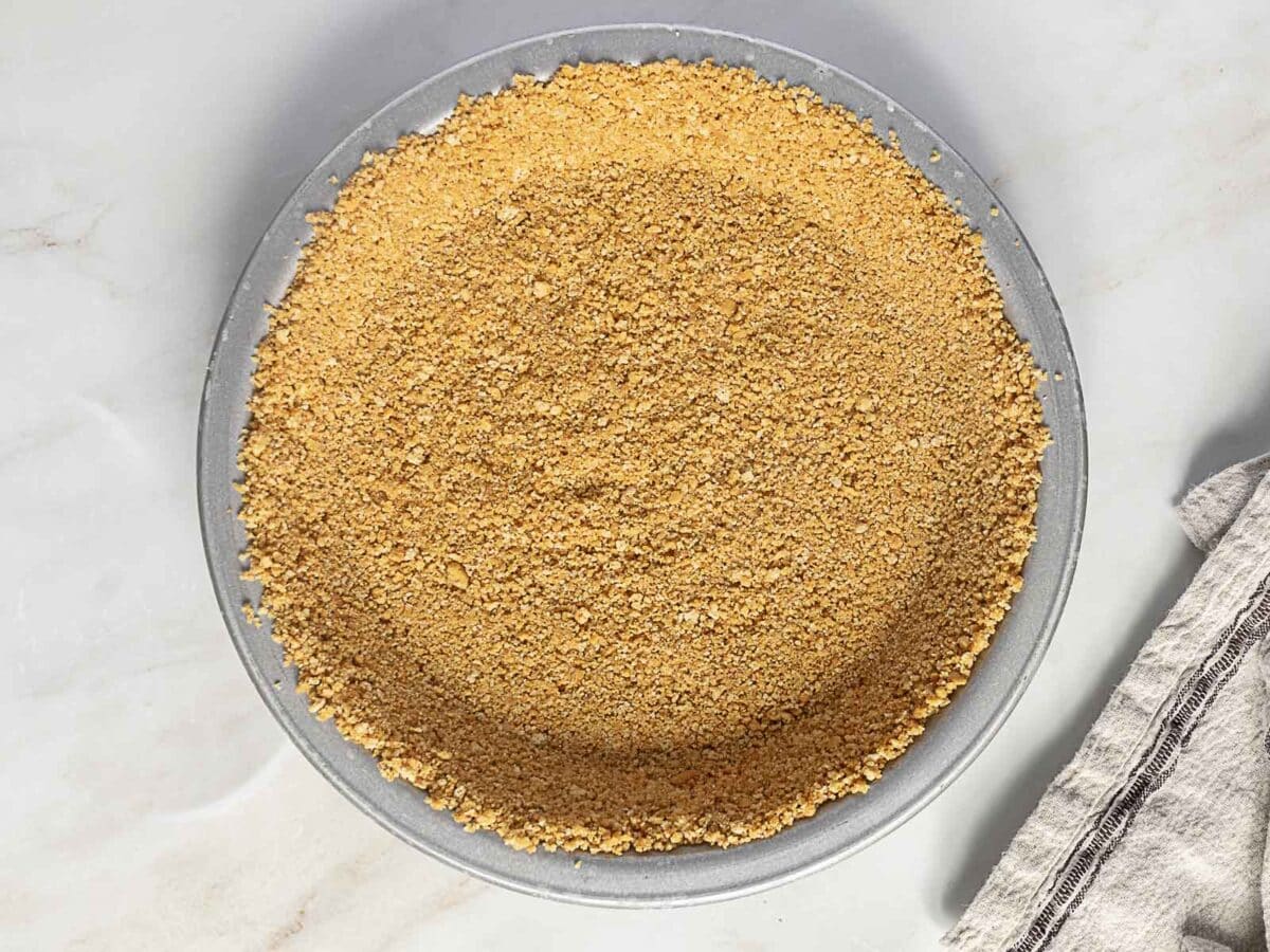 A pie dish filled with a pressed graham cracker crumb crust, viewed from above on a light surface next to a striped towel.