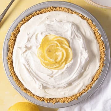 A lemon cream pie with a graham cracker crust, topped with whipped cream and lemon slices, sits on a light surface next to a whole lemon.