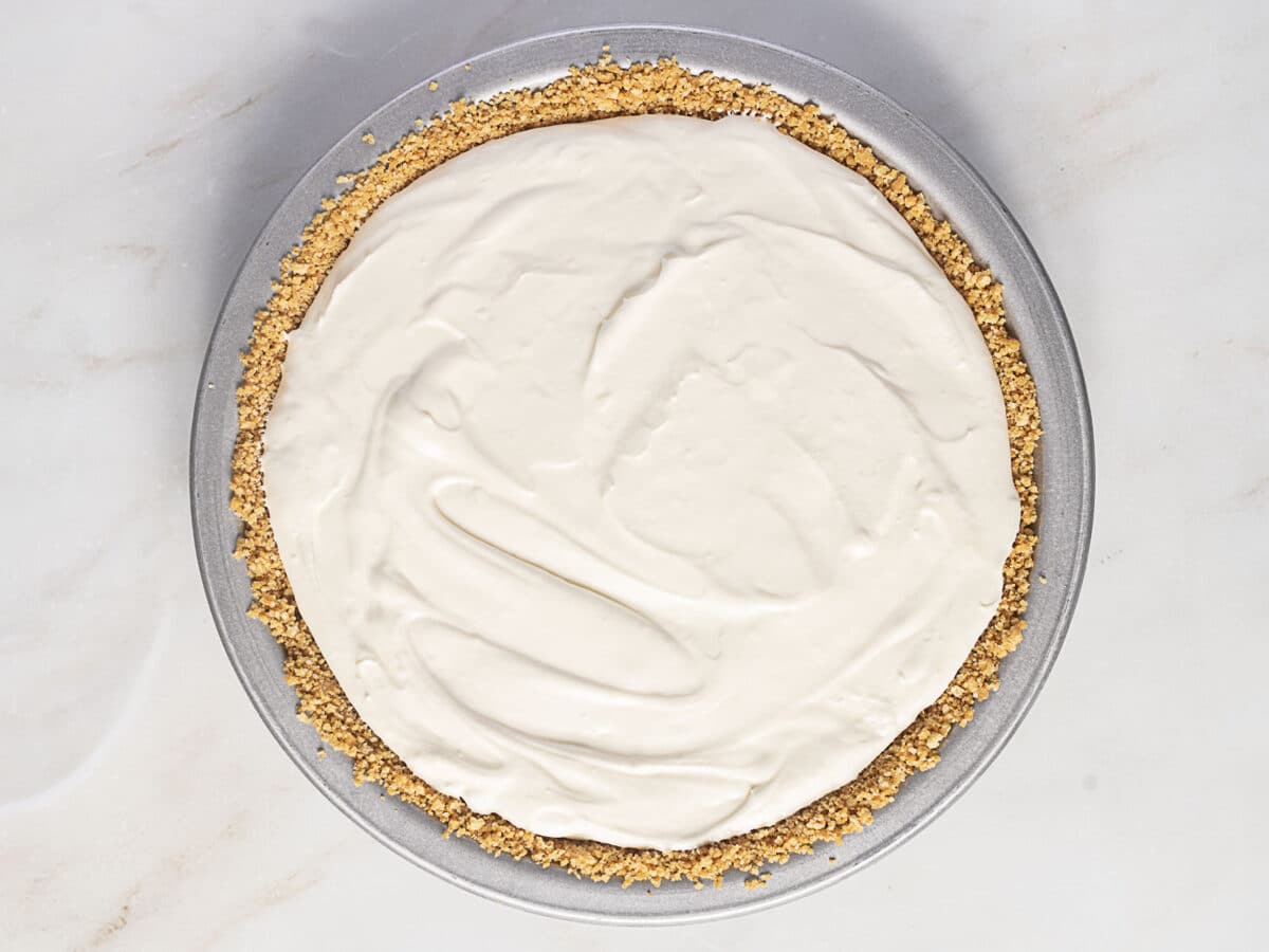 A cheesecake with a graham cracker crust is shown in a round metal pan on a white surface. The top is smooth and creamy, with no toppings.