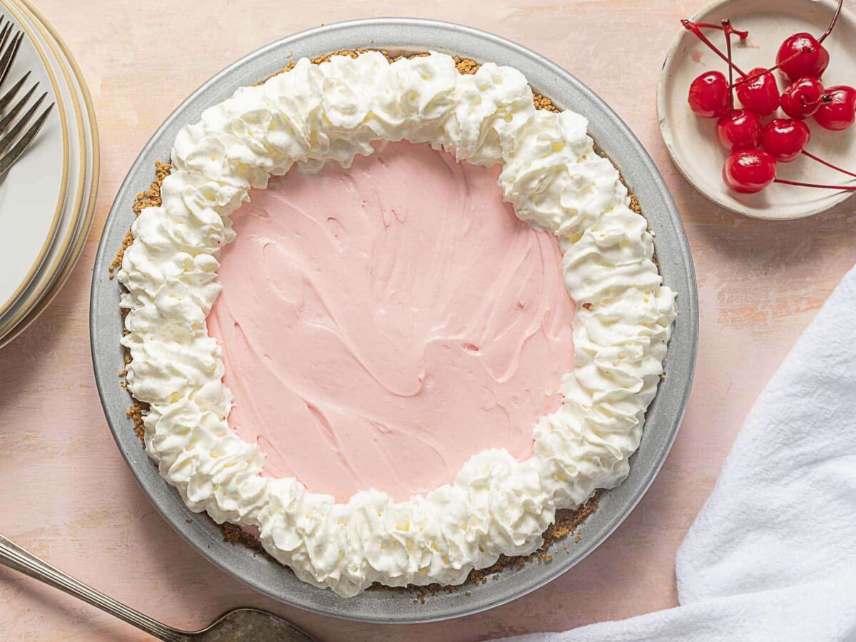 A graham cracker crust pie with pink filling and a border of whipped cream, next to a dish of cherries, plates, and a white napkin.