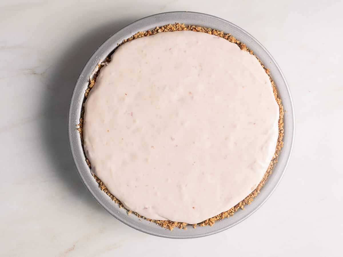 A round pie with a smooth, pale pink filling in a graham cracker crust sits in a metal pie pan on a white surface.