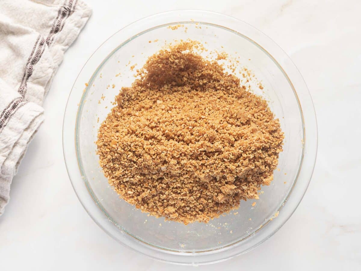 A glass bowl filled with a crumbly mixture, likely graham cracker crust, sits on a light-colored surface next to a folded kitchen towel.