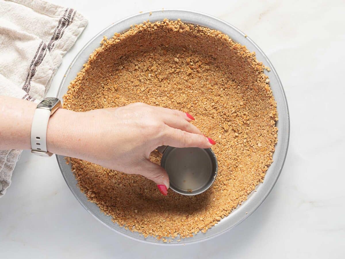 A hand presses a round metal measuring cup into a pie pan filled with crumb crust mixture on a white surface.