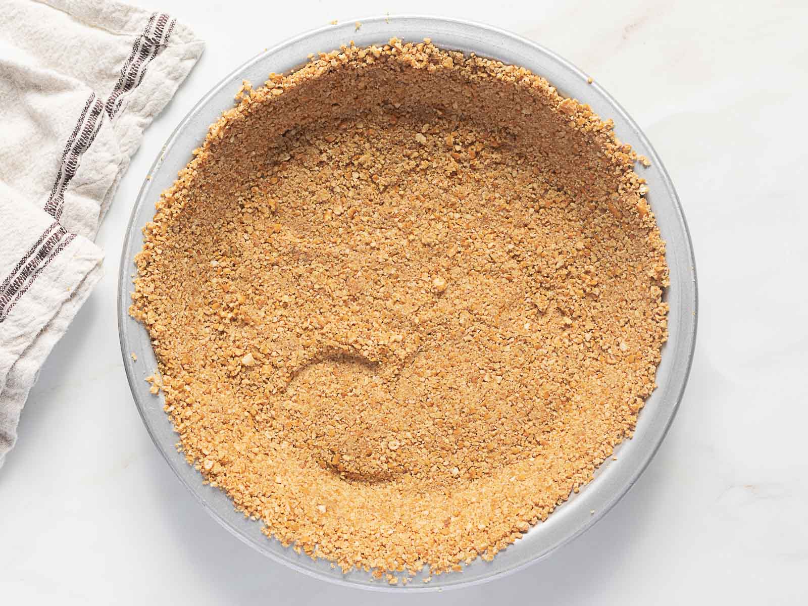 A pie pan filled with a pressed graham cracker crumb crust, with an area near the bottom showing a swipe mark. A striped cloth is partially visible to the side.