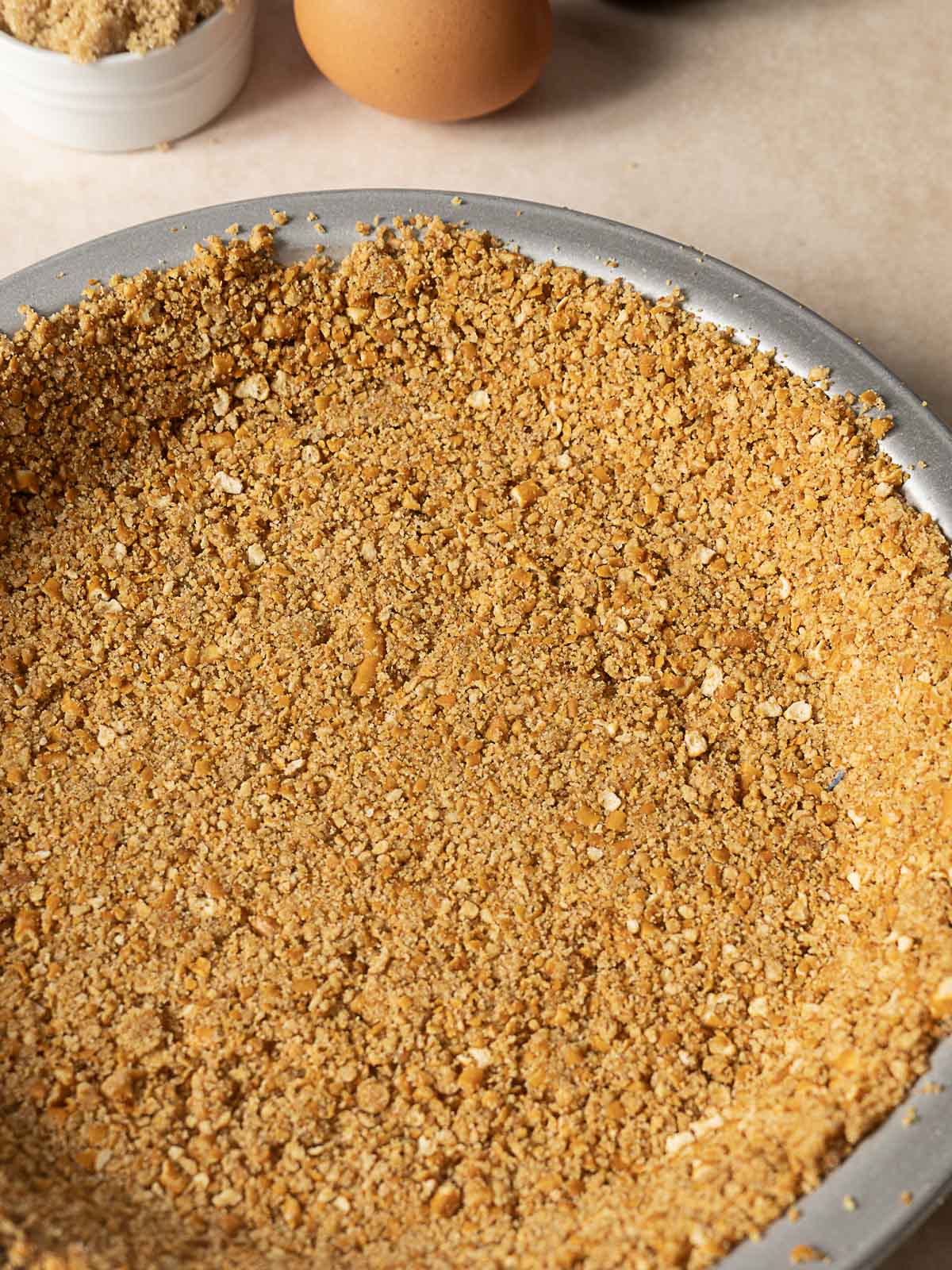 A close-up of a pie dish filled with a pressed graham cracker crumb crust, ready to be baked. An egg and a small bowl of brown sugar are visible in the background.