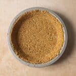 A round metal pie dish filled with a pressed, crumbly graham cracker crust on a light beige surface.
