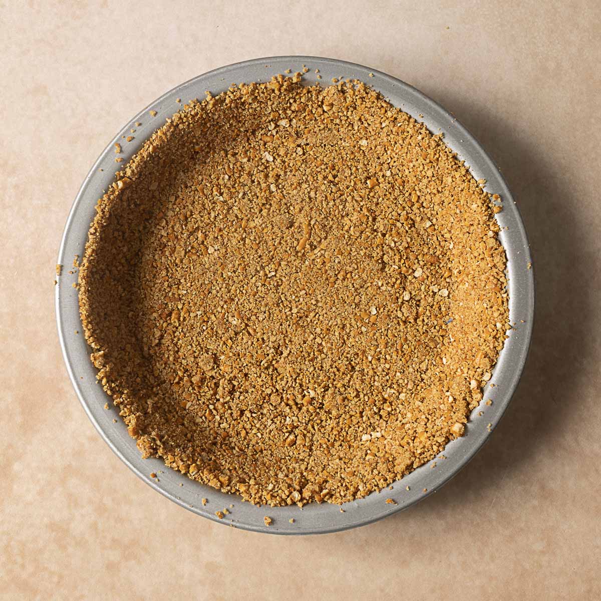 A round metal pie dish filled with a pressed, crumbly graham cracker crust on a light beige surface.