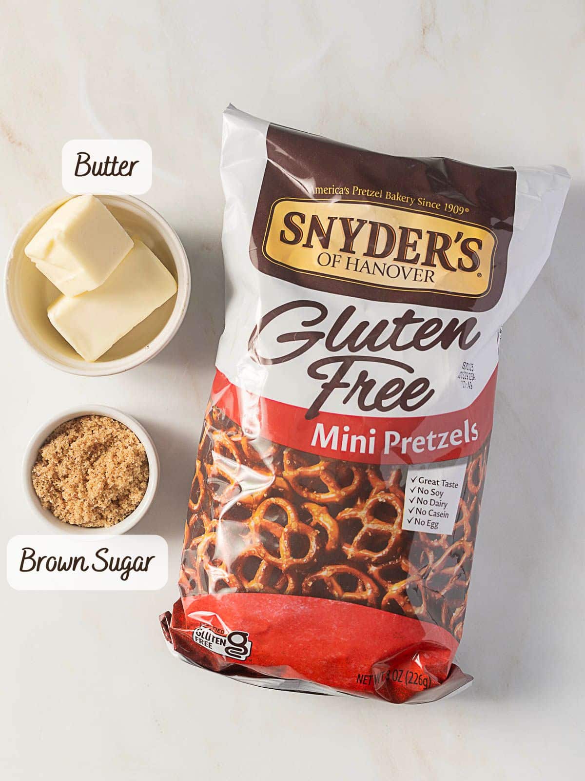 A bag of Snyder’s of Hanover gluten free mini pretzels next to small bowls of butter and brown sugar on a white surface.