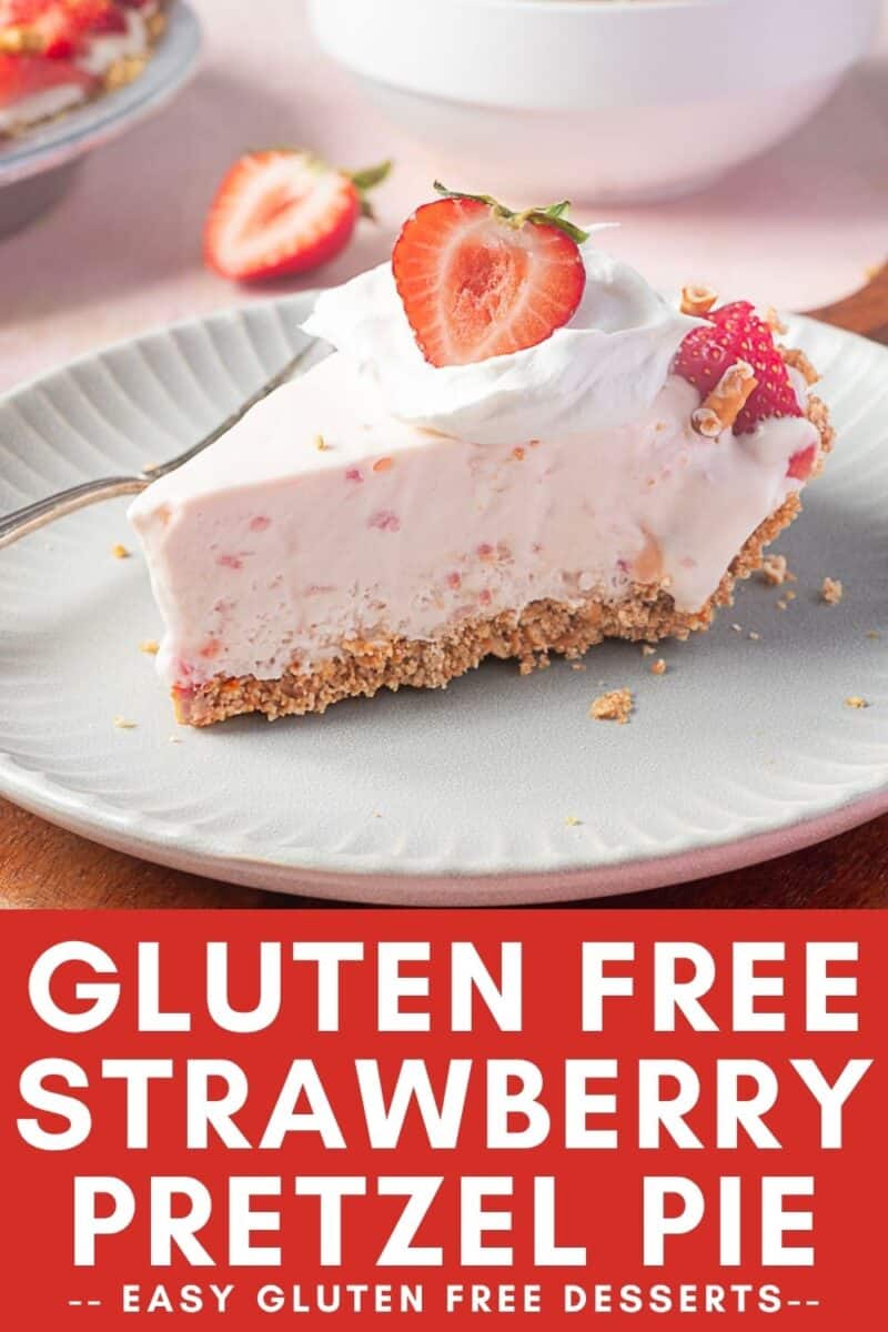 A slice of gluten free strawberry pretzel pie topped with whipped cream and a strawberry half sits on a plate, with another slice and strawberries in the background.