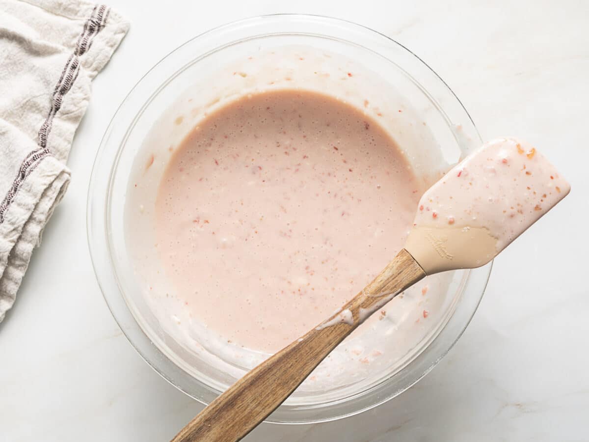 A glass bowl filled with a light pink, speckled batter sits on a white surface next to a beige cloth; a spatula rests on the bowl’s edge.