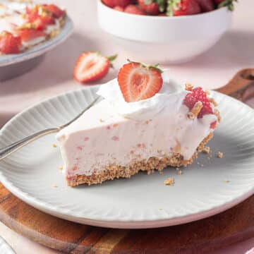 A slice of strawberry cheesecake on a plate with whipped cream and a fresh strawberry on top, with a fork inserted, and a bowl of strawberries in the background.