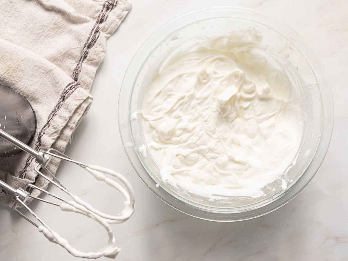 A glass bowl filled with whipped cream sits on a marble surface next to a towel and a handheld electric mixer with some cream on the beaters.