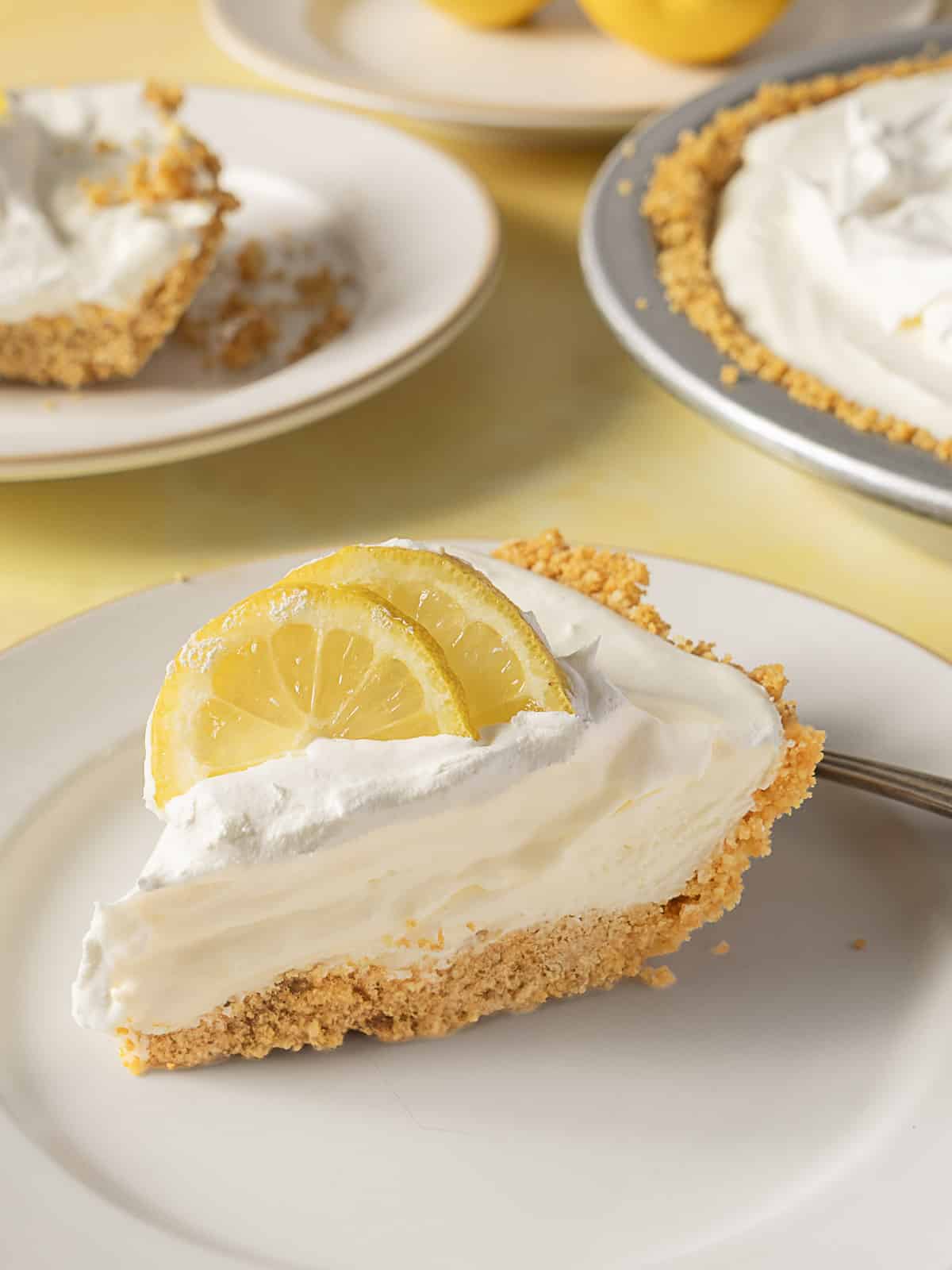 A slice of lemon cream pie with whipped topping and two lemon slices sits on a white plate; the whole pie and another plated slice are visible in the background.