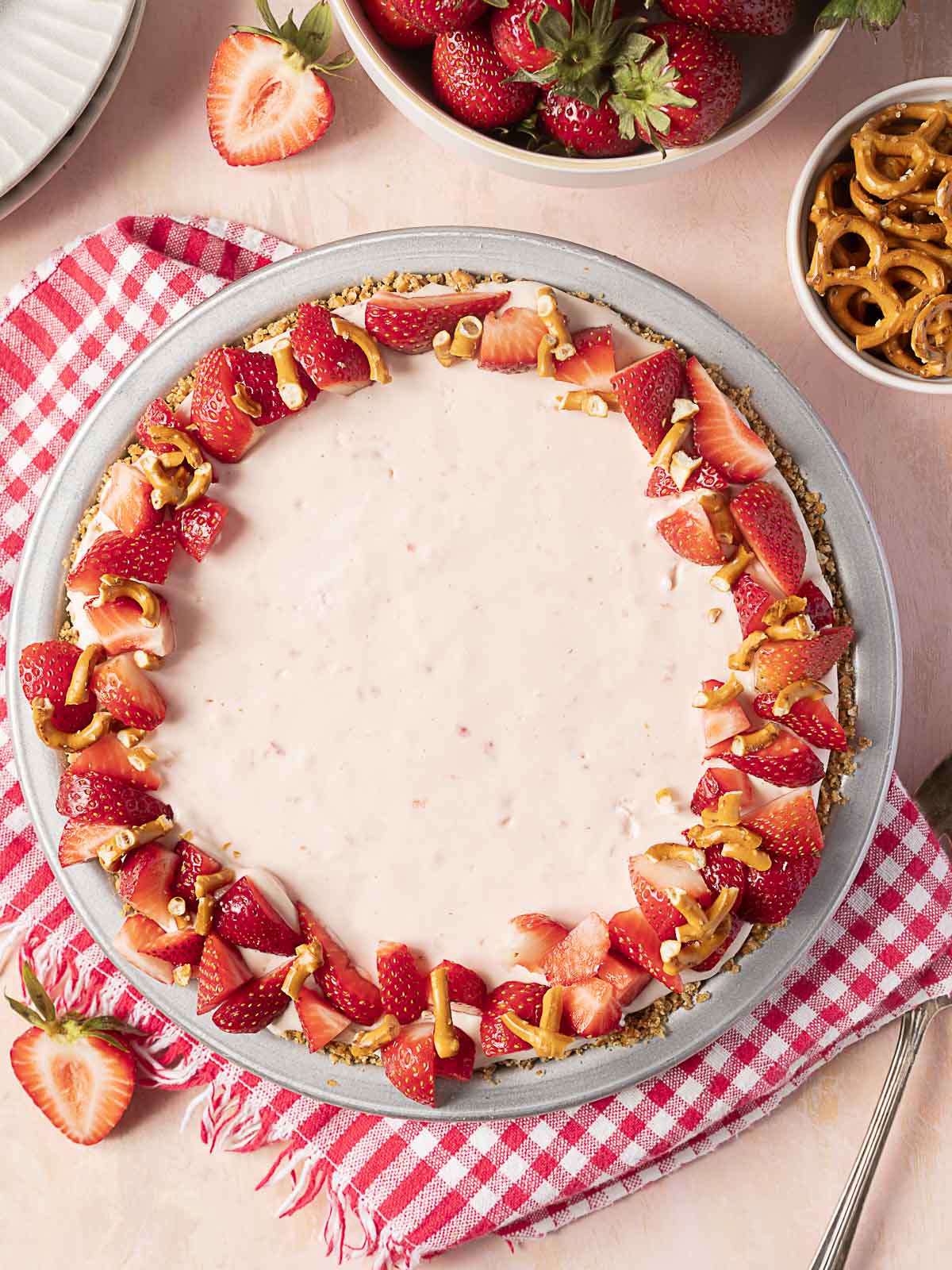 A strawberry cheesecake topped with fresh strawberries and pretzel pieces sits on a plate beside a red checkered napkin, with bowls of strawberries and pretzels nearby.