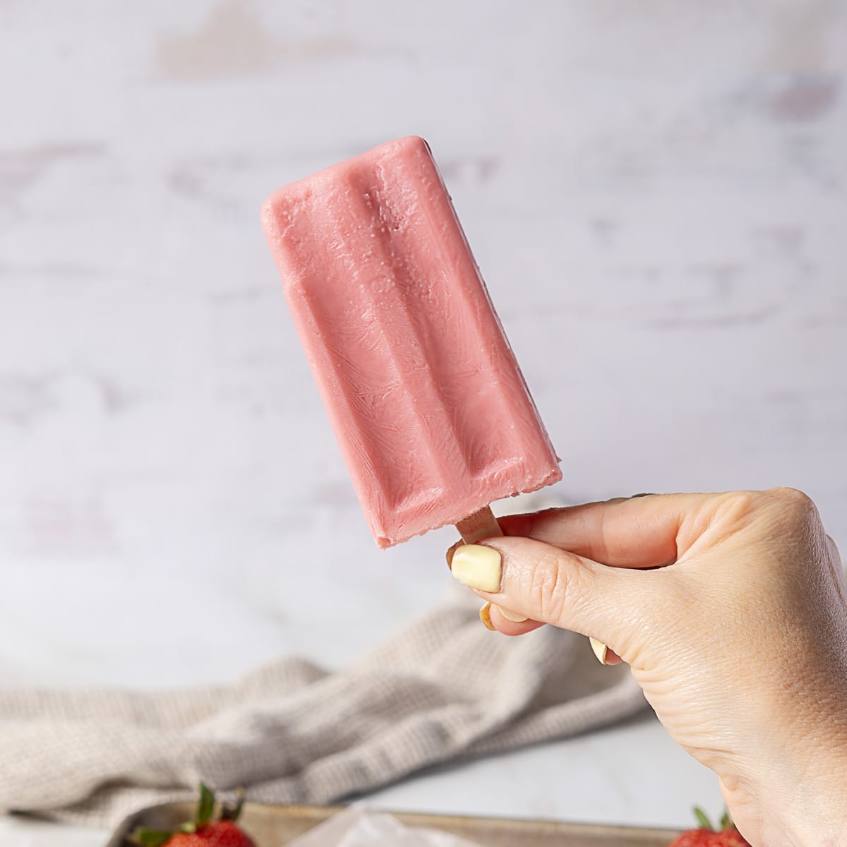 A hand holding a pink popsicle on a wooden stick against a light background.
