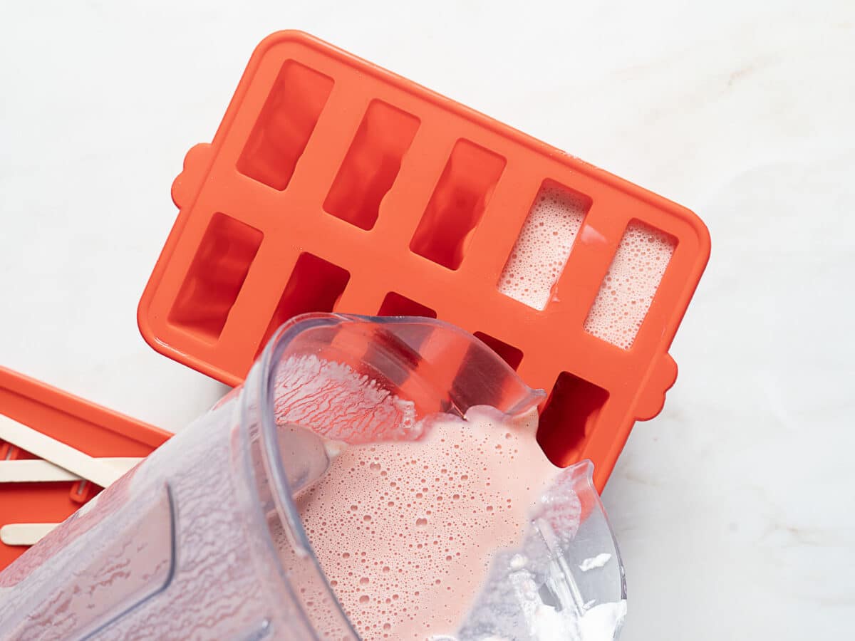 A blender pours pink smoothie mixture into a red popsicle mold tray on a white surface, with wooden sticks beside it.