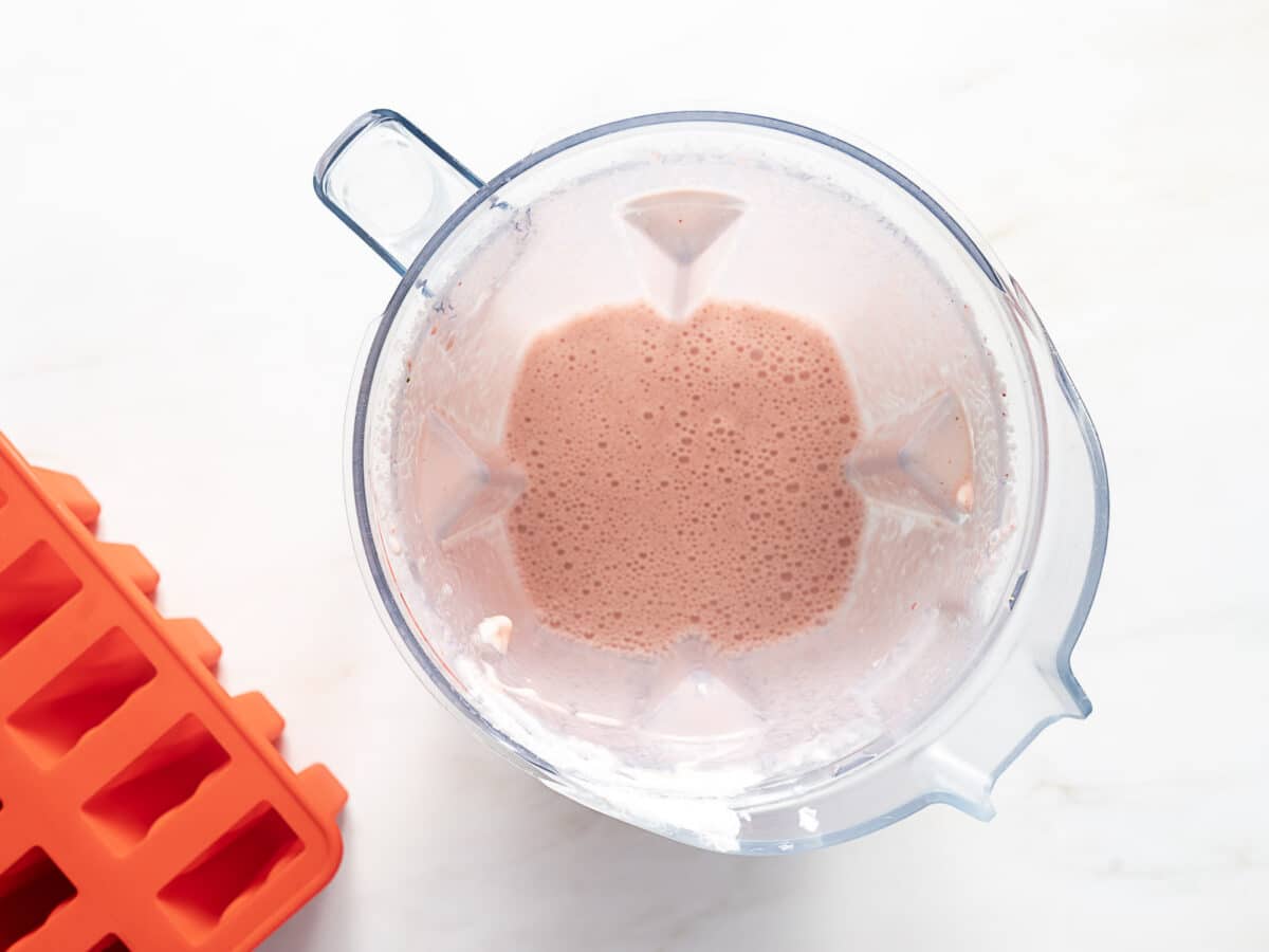 A blender containing a pink smoothie mixture sits beside a red ice cube tray on a white surface.
