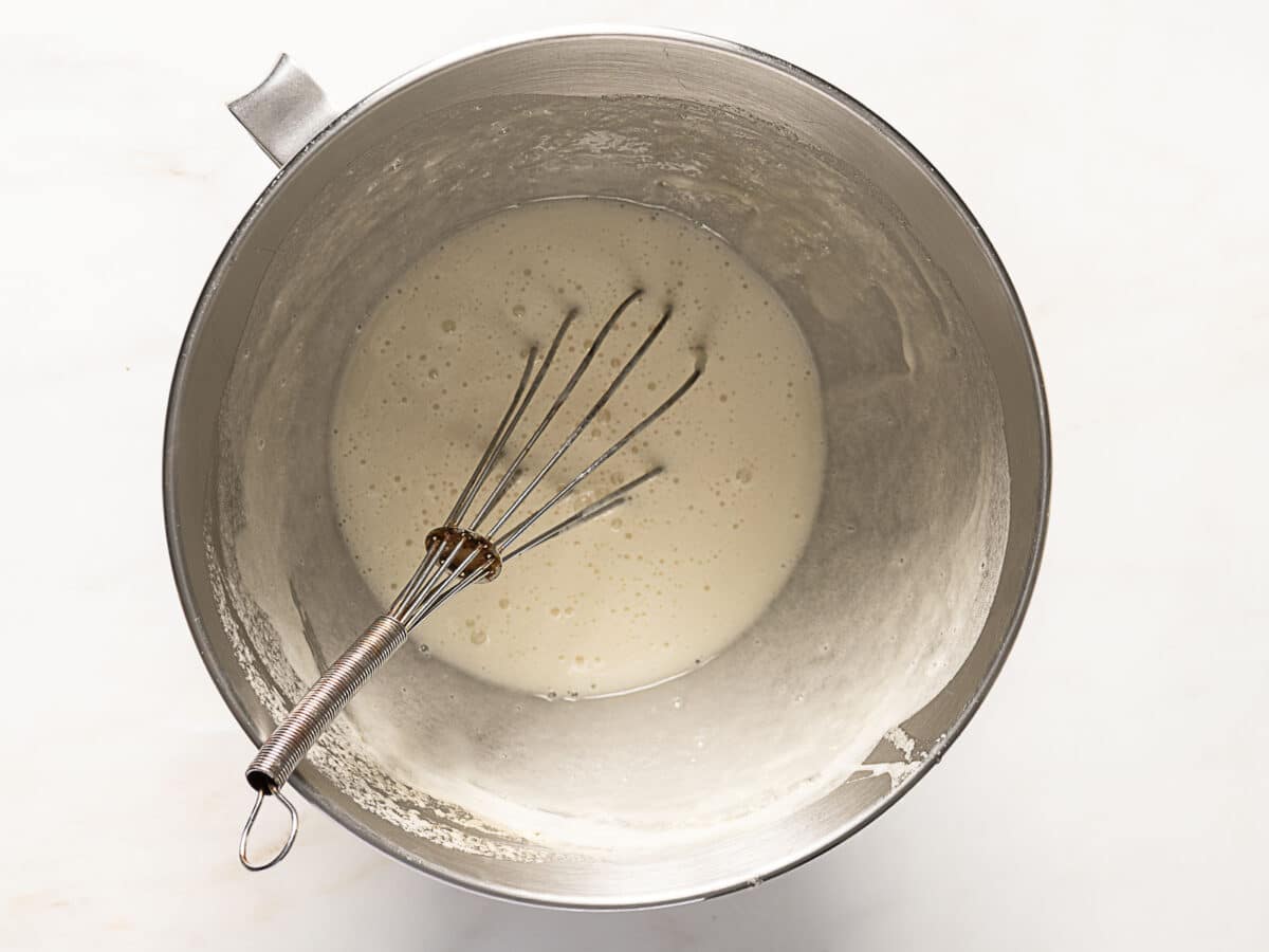 A metal whisk rests in a stainless steel mixing bowl containing a smooth, light batter.