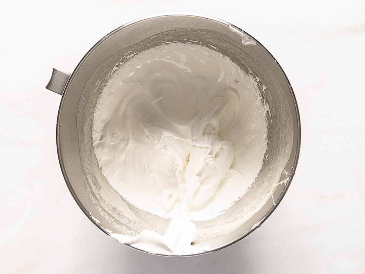 A metal mixing bowl filled with a thick, glossy white mixture that appears to be whipped cream or meringue on a white surface.
