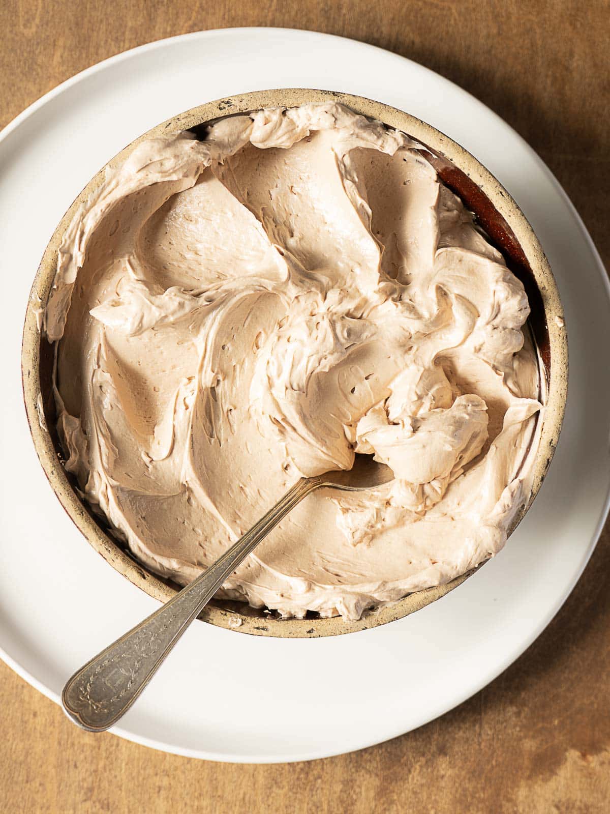 A ceramic bowl filled with whipped chocolate frosting, with a spoon resting inside, placed on a white plate on a wooden surface.