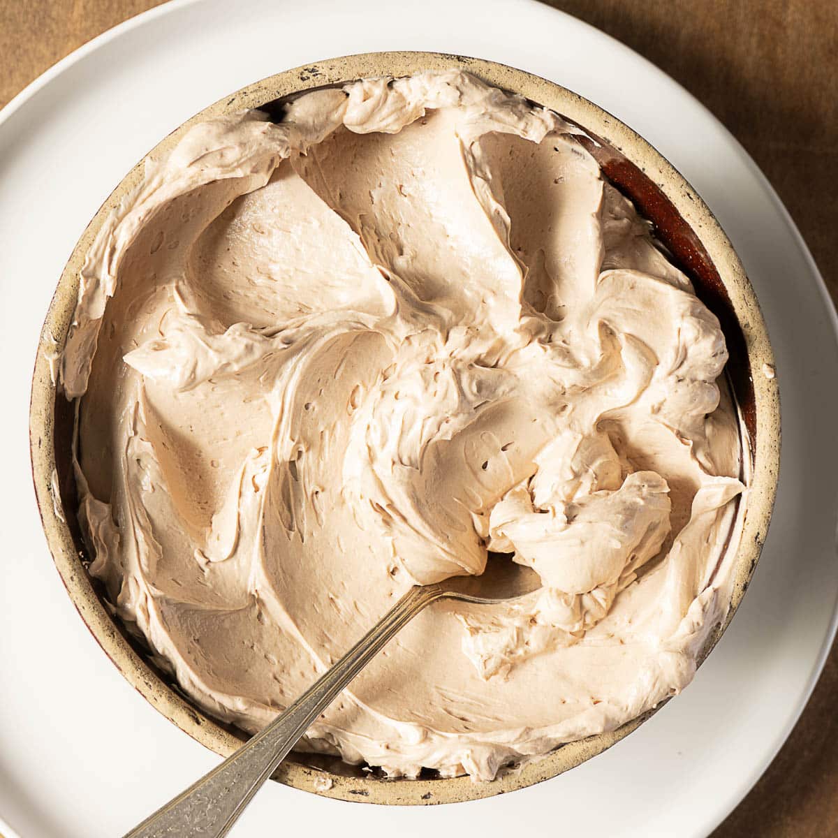 A bowl of light brown whipped frosting with a spoon in it, placed on a white plate.