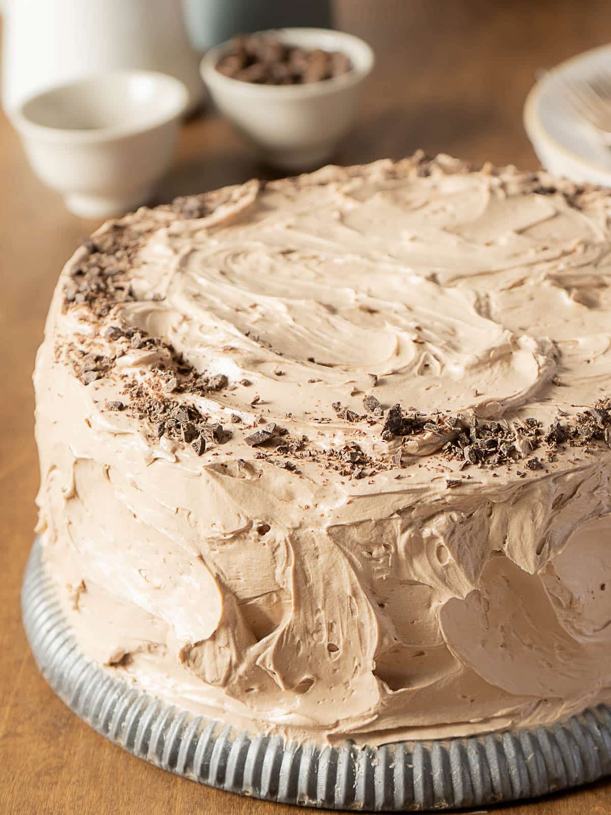 A round chocolate cake with swirled chocolate frosting and chocolate shavings on top, displayed on a metal platter.