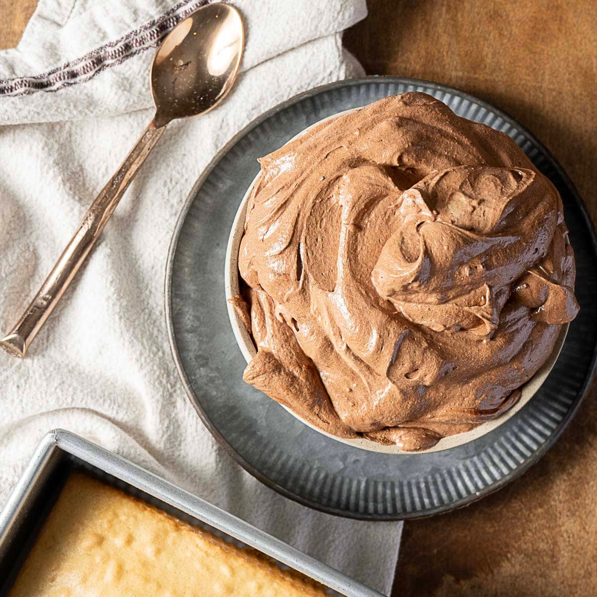 A bowl of chocolate frosting sits on a plate next to a spoon, a white cloth, and a partially visible baked cake in a metal pan.
