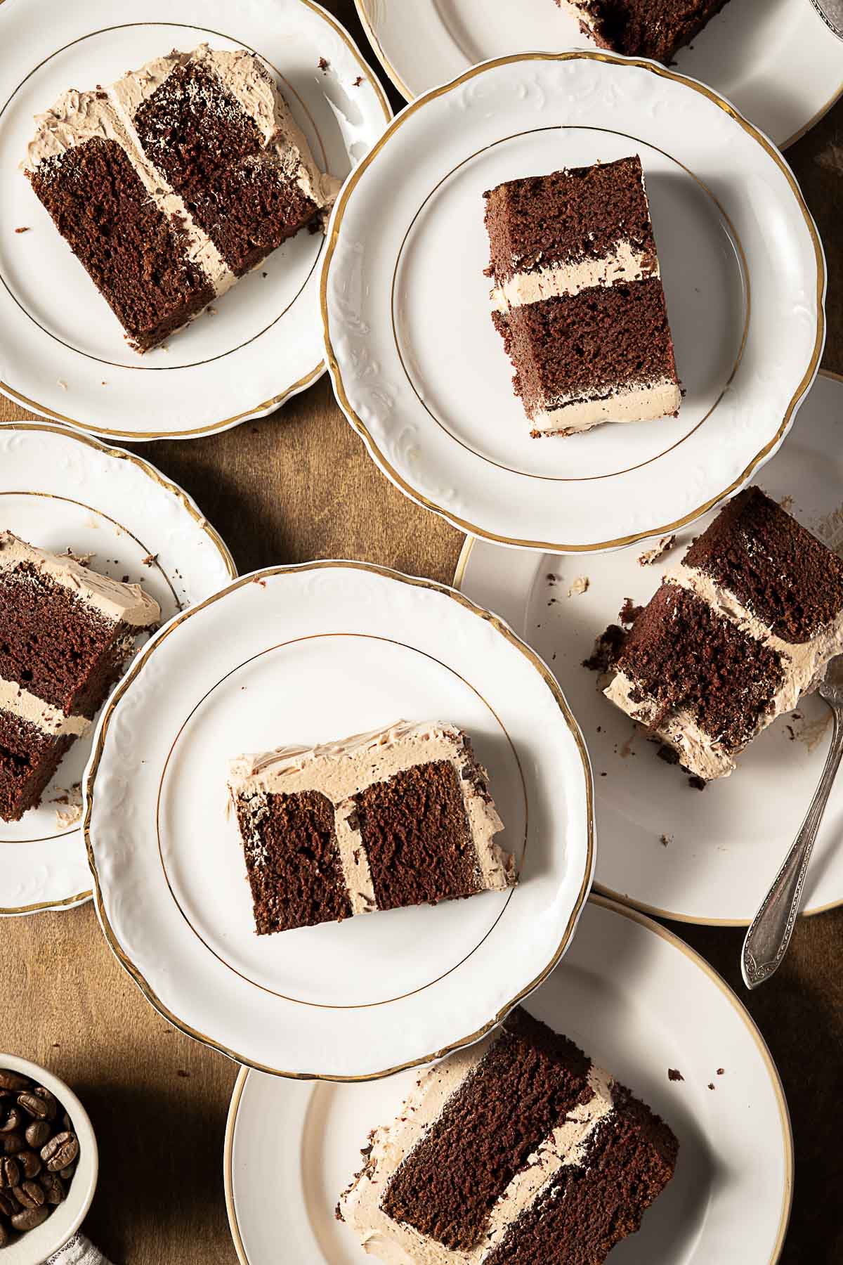 Several white plates, each with a slice of chocolate layer cake with light brown frosting, are arranged on a table. A spoon and a small bowl of coffee beans are also visible.